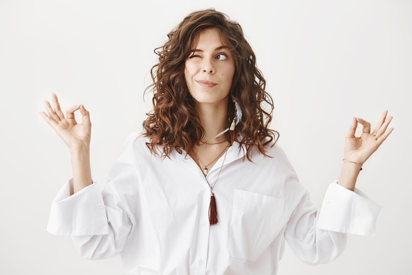 Studio portrait of charming positive caucasian female businesswoman trying to relax while meditating, standing with lifted hands and zen signs over gray background, peeking and looking aside playfully